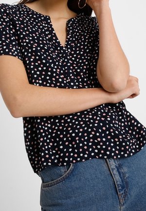 Femme portant un chemisier noir à manches courtes avec un motif de pois roses et blancs et un jean bleu, bras croisés, une main touchant le visage.