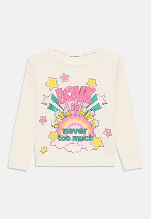 Chemise à manches longues crème avec texte coloré pailleté "Love is never too much" entouré d'étoiles, de nuages et de formes d'arc-en-ciel à l'avant.