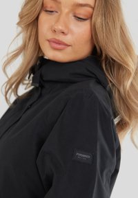 Chaqueta negra hecha de un material texturizado, con un cuello alto, cierre de botones y una etiqueta de marca visible en la manga izquierda.