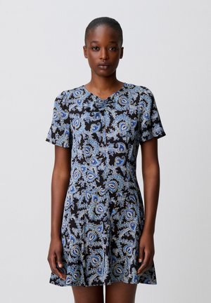 Donna che indossa un vestito nero a maniche corte con motivi paisley blu e bianchi, in piedi su uno sfondo grigio chiaro uniforme.
