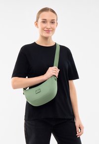 Borsa a spalla in tessuto verde con forma arrotondata e tracolla regolabile. Presenta un piccolo tag con logo sulla parte anteriore e una texture liscia.