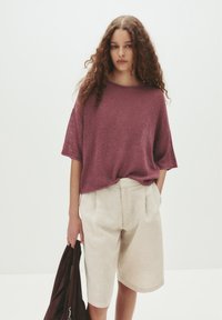 Burgunderfarbenes kurzärmeliges Strickoberteil, kombiniert mit hellbeigen Leinen-Shorts. Das Outfit umfasst eine braune Jacke, die in einer Hand gehalten wird.