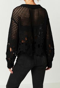 Czarny sweter z szydełkowanego splotu o otwartym wzorze z kwiatowymi akcentami, długimi rękawami i krótkim dołem. Noszony na ciemnych dżinsach.