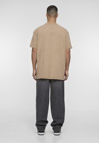 Camiseta oversize color caqui con mangas cortas, cuello redondo y detalles de costura visibles, combinada con pantalones oscuros de corte amplio y zapatillas.