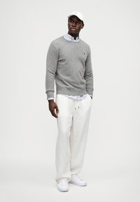 Γκρι πουλόβερ με στρογγυλή λαιμόκοψη, μπλε καρό πουκάμισο από κάτω, λευκό παντελόνι από λινό και λευκά αθλητικά παπούτσια. Λευκό καπέλο με λογότυπο στην κορυφή.