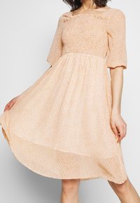 Žena oblečená v midi šatech světle broskvové barvy s drobným květinovým vzorem, krátkými nafouklými rukávy a čtvercovým výstřihem, držící lem šatů.