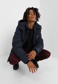 Marineblå pufferjakke med hætte, lynlås, quiltet mønster, parret med en sort sweater og bordeauxfarvede bukser. Sorte sneakers fuldender looket.
