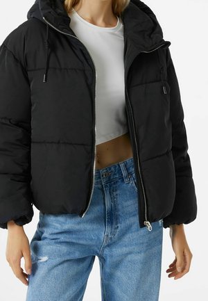 Femme portant une doudoune noire à capuche par-dessus un t-shirt court blanc et un jean bleu taille haute sur fond blanc.