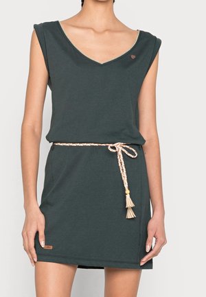 Hihaton tummanvihreä mekko, jossa on V-pääntie ja solmittu beige punottu köysivyö vyötäröllä, päällä seisova henkilö, jonka kädet ovat rentoina.