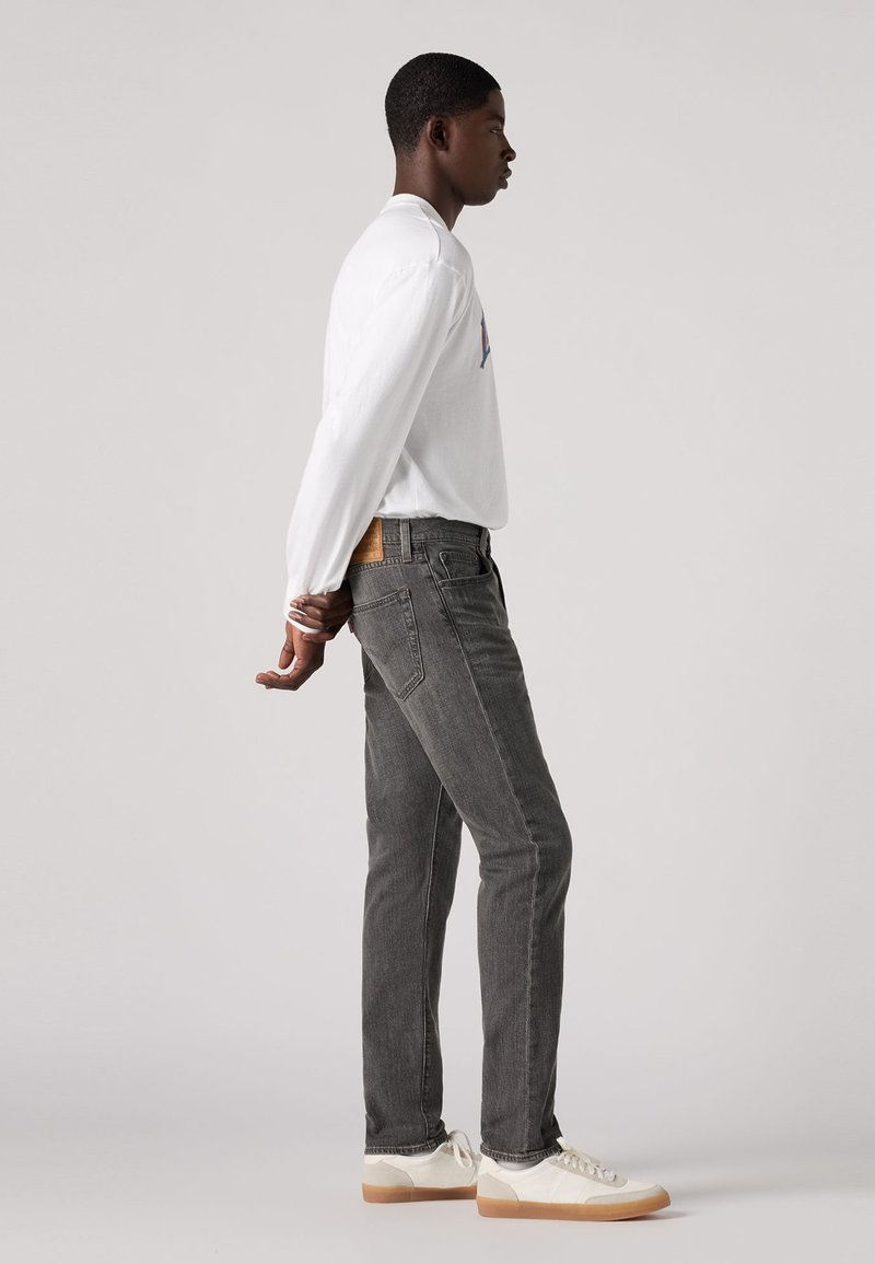 Jean en denim gris avec une coupe droite, associé à une chemise blanche à manches longues et des baskets de couleur claire. Détail notable sur la poche arrière visible.