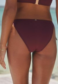 Bikini w kolorze bordo z gładką teksturą, niskim stanem i minimalnym wycięciem po bokach, z małym akcentem logo w pobliżu pasa.