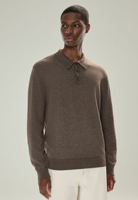 Brauner langärmeliger Polo-Pullover mit geripptem Kragen, einer Knopfleiste mit drei Knöpfen und strukturiertem Stoff, kombiniert mit hellen Hosen.