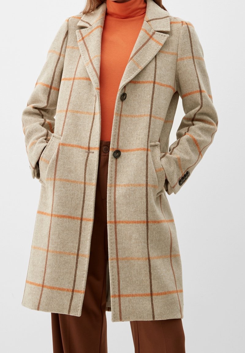 Abrigo de lana beige con patrón de cuadros en naranja y marrón, solapas con muesca, cierre de dos botones y bolsillos laterales. Llevado sobre un suéter de cuello alto naranja.