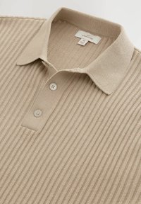 Beige ribbet polo-shirt med krave, tre-knaps placket og subtil tekstur. Tag viser størrelse M og mærkenavn.