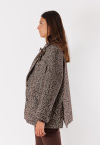 Cappotto marrone a motivo herringbone, tessuto testurizzato, taglio oversize, colletto prominente, chiusura con grande bottone e dettaglio a fiocco su una spalla.