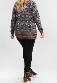 Langärmlige, gemusterte Bluse in Schwarz, Beige und Burgunderrot, kombiniert mit schwarzen Skinny-Jeans und Ankle-Boots mit Holzabsatz.