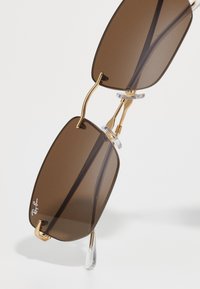 Hnedé obdĺžnikové slnečné okuliare Ray-Ban so zlatými kovovými rámami a priehľadnými nosovými podložkami na bielom pozadí.