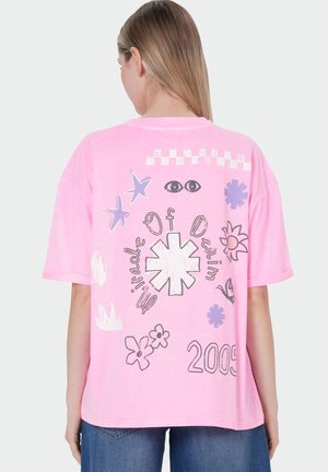 Personne blonde portant un T-shirt rose ample avec des motifs floraux et d’yeux violets et blancs, du texte et le chiffre 2009 au dos.