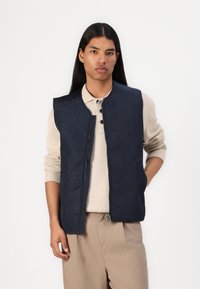 Marineblauwe gewatteerde vest met geometrisch patroon, ritsluiting en ronde halslijn over een beige trui. Model houdt een hand op de taille.