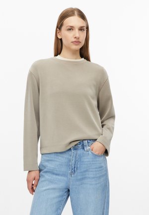 Junge Frau mit glatten Haaren, die ein beiges Langarmshirt und hellblaue Jeans trägt, steht mit einer Hand in der Tasche.