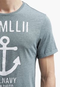 Gray short-sleeve T-shirt made of soft fabric, featuring a white anchor design and text. The sleeves are rolled up, showing a smooth texture.