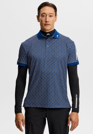 Mand iført blå poloshirt med geometrisk mønster over sorte lange ærmer og sorte bukser, stående mod en ensfarvet hvid baggrund.