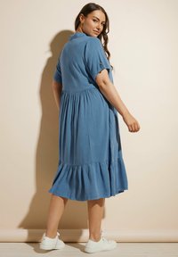 Abito azzurro chiaro con maniche corte, orlo arricciato e vita arricciata. Abbinato a sneakers bianche su uno sfondo neutro.