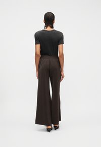 Άτομο με πλεξούδες, που φοράει ένα ξεθωριασμένο μαύρο μπλουζάκι, φαρδιά καφέ σκούρα παντελόνια και μαύρα παπούτσια με τακούνι, όρθιο με την πλάτη προς το μέρος μας σε λευκό φόντο.