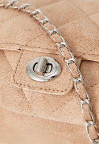 Beige tekoturkistakangas käsilaukku, jossa on poltettu muotoilu, hopeiset metalliosat ja ketjuhihna. Varustettu pyöreällä painonappilukolla.