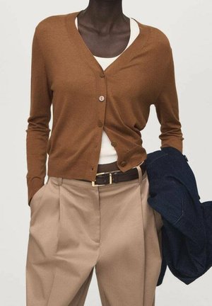 Modelka nosi brązowy sweter na guziki na biały top, beżowe plisowane spodnie z paskiem, trzyma ciemnoniebieską kurtkę w jednej ręce.