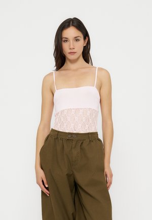 Mujer con una blusa de encaje blanca con tirantes de espagueti y pantalones sueltos de talle alto de color verde oliva frente a un fondo liso.