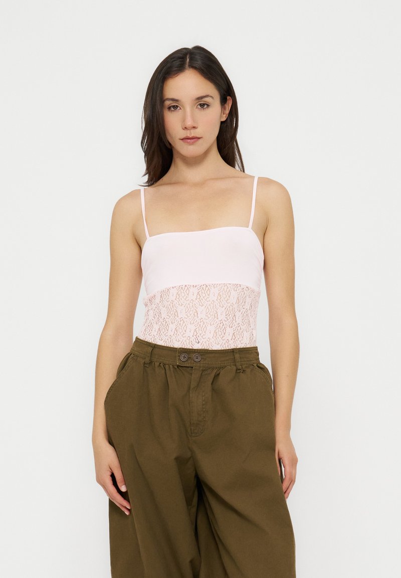 Femme portant un haut caraco en dentelle blanche avec des bretelles spaghetti et un pantalon ample taille haute vert olive sur un fond uni.