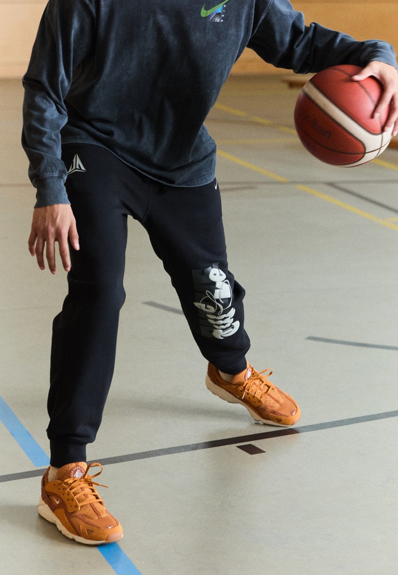 Kosárlabdázó dribblingozik. Szürke hosszúujjú pólót és fekete melegítőnadrágot visel, fehér nyomattal. Narancssárga sportcipői perforált részletekkel rendelkeznek.