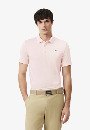 Uomo che indossa una polo rosa chiaro con un logo di coccodrillo verde e pantaloni beige con una cintura abbinata, in piedi con una mano in tasca.