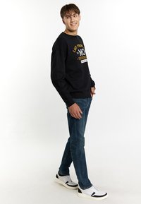 Felpa nera con grafica testuale gialla e bianca, jeans blu e sneakers bianche con accenti neri; modello in piedi di lato.
