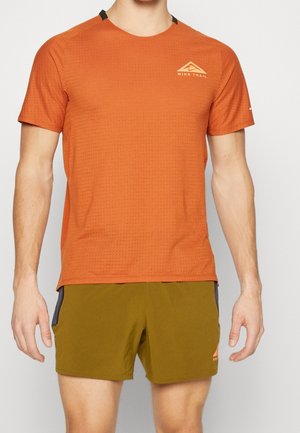 Mężczyzna noszący pomarańczową koszulkę Nike Trail z krótkim rękawem i oliwkowe krótkie spodenki, stojący na tle jasnego, jednolitego tła.