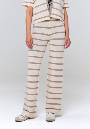 Femme portant un pantalon beige et marron clair à rayures horizontales et un haut assorti, associé à des chaussures blanches slip-on avec des motifs découpés.