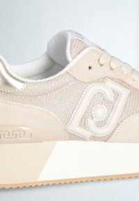 Sneaker beige con tessuto testurizzato e dettagli in camoscio. Presenta un logo metallico, lacci color crema e una suola bianca con fondo marrone chiaro.