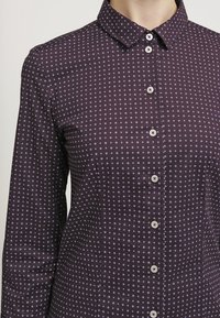 Chemise à manches longues en tissu violet foncé, avec un motif à pois blancs, un col structuré et des boutons en argent. Texture lisse.