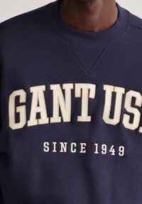 Námořnická mikina s vyvýšeným béžovým nápisem "GANT US" a "SINCE 1949." Materiál je silný a strukturovaný, s kruhovým výstřihem a žebrovanými manžetami.