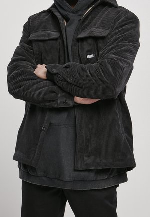 Personne portant une veste en velours côtelé noire par-dessus un sweat à capuche noir, bras croisés, avec l'étiquette de la veste indiquant Urban Classics.