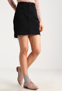 Femme portant une mini-jupe en denim noir à ourlet effiloché, un haut à manches longues rose clair, des chaussettes grises et des chaussures roses à lacets, debout devant un fond uni.