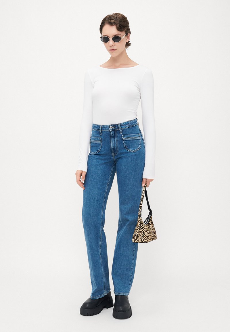 Woman in white long-sleeve top, blue high-waist jeans, black platform boots, sunglasses, holding small zebra-print handbag.
