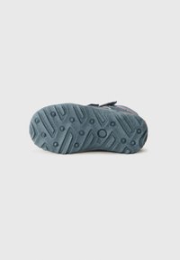 Sola de sapato em plástico azul com um padrão texturizado, apresentando sulcos profundos e aderências circulares para tração. Detalhe da fita de velcro visível.