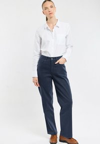 Chemise blanche à manches longues avec poche, associée à un pantalon ample bleu marine et des chaussures marron. Tissu lisse et design décontracté.