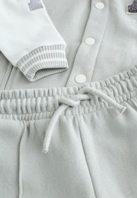 Felpa grigio chiaro con bottoni bianchi e polsini a righe, abbinata a joggers grigio chiaro coordinati con coulisse e vita arricciata.