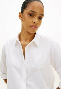 Mujer con el cabello peinado hacia atrás, vistiendo una camisa blanca con botones y un pequeño logo bordado en el pecho, mirando ligeramente hacia la cámara.