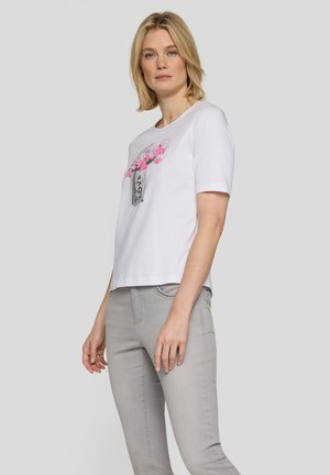 Woman standing wearing white short-sleeve shirt with pink floral vase design and light gray pants against plain light gray background.