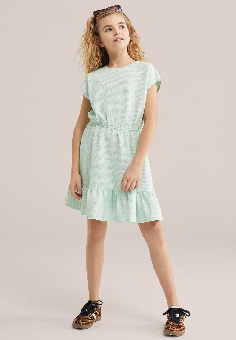 Jeune fille aux cheveux bouclés portant une robe texturée vert clair et des baskets à imprimé léopard, debout les mains relâchées et regardant vers le haut.
