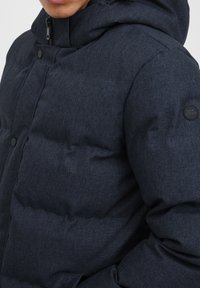 Mörkblå pufferjacka med huva. Har en texturerad, vadderad design och svarta tryckknappar. Logotypapplikation på vänster ärm.